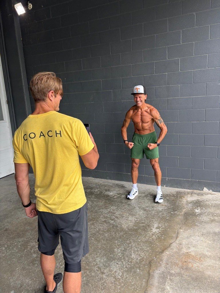 Dai Manuel, seen from behind in a yellow COACH t-shirt, photographing a lean older client flexing against a grey wall